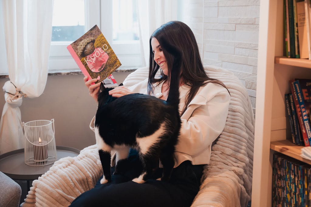 Une cliente sourit en lisant un livre dans un fauteuil moelleux, un chat tuxedo sur ses genoux — l'ambiance ronronthérapie chez Cats & Chill